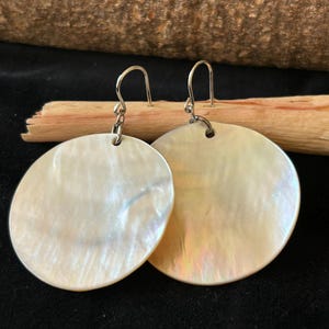 May include: A pair of iridescent, round earrings with silver-colored hooks. The earrings have a pearlescent sheen, reflecting light with hints of pink and blue. The earrings are displayed against a dark background with a wooden accent.