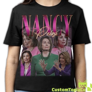 May include: Black t-shirt featuring a graphic of Nancy Pelosi with the name "NANCY" in a pink and gold font. The design includes multiple images of Nancy Pelosi. The CustomTopiaCo logo is at the bottom.