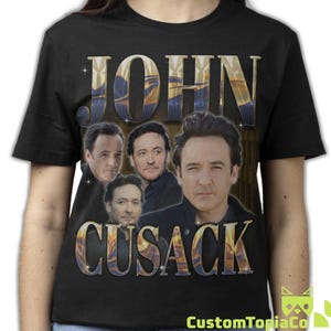May include: A black short-sleeve t-shirt featuring a graphic print of actor John Cusack in four different poses. The name JOHN CUSACK is printed in large, metallic-toned, stylized lettering above and below the images, accented with small star icons.