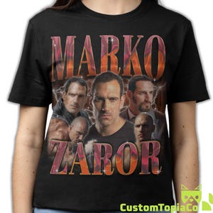 May include: Black t-shirt featuring a collage of images of a man with the name "MARKO ZABOR" in a gradient orange and red font. The shirt has a vintage, retro design with a CustomTopiaCo logo.