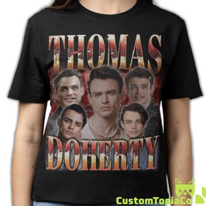 May include: Black t-shirt featuring a vintage-style design with the name "THOMAS DOHERTY" in a distressed, orange and red font. The shirt showcases multiple images of a man. The CustomTopiaCo logo is visible at the bottom right.