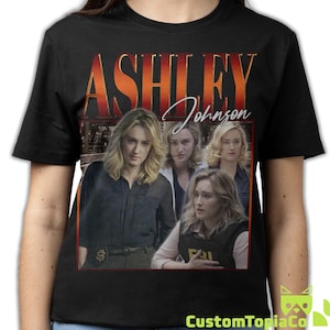 Op de afbeelding: Een zwart t-shirt met korte mouwen en een grafische print van actrice Ashley Johnson. Het ontwerp bevat de naam ASHLEY in grote rode en oranje letters, de naam Johnson in wit handschrift en vier verschillende afbeeldingen van de actrice.