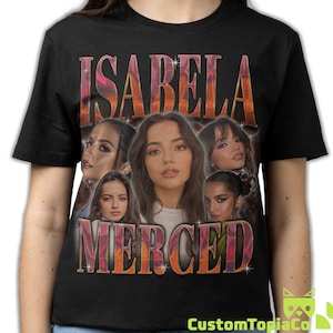 May include: Black t-shirt with the name "ISABELA MERCED" in a retro style, with a portrait of the actress. The design includes multiple images of the actress. The shirt has a crew neck.