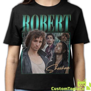 May include: A black short-sleeved t-shirt featuring a graphic print of actor Robert Sheehan. The design includes the name ROBERT in large teal letters at the top and the name Sheehan in a gold script font at the bottom, overlaying multiple images.