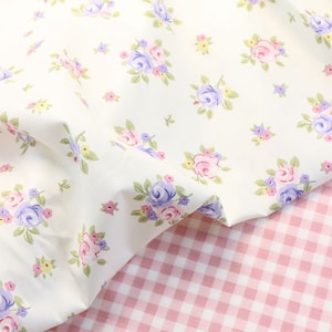 May include: Cream-coloured fabric adorned with a floral design of pink, purple, and blue roses, complemented by green foliage. Beneath, a pink and white gingham pattern provides a contrasting backdrop, enhancing the visual texture.