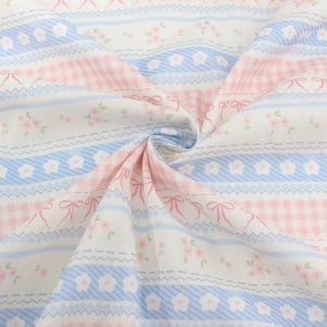 May include: Close-up of a fabric with a repeating pattern. Pink gingham, light blue stripes, and white sections are visible. The white sections feature small pink flowers and bows. The fabric is folded, showing the repeating design.