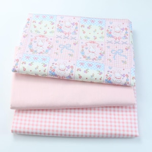 May include: Three stacks of fabric in shades of pink. The top layer features a patchwork design with bunnies, flowers, and bows. The middle layer is a solid pink, and the bottom layer has a pink and white gingham pattern.