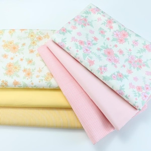 May include: A stack of fabric samples featuring floral, striped, and solid patterns. The fabrics include a light yellow with orange flowers, a light green with pink flowers, a pink and white striped fabric, a solid pink fabric, and a yellow and white striped fabric.