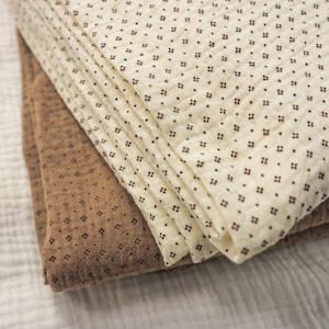 May include: Folded cream and brown fabric with a repeating dark brown square pattern. The fabric appears to be soft and textured, possibly cotton or linen. The layers are neatly stacked, showcasing the different colours and patterns.