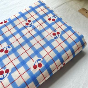 May include: A folded fabric featuring a blue and white gingham pattern, with a red grid and cherry motifs. The cherries are red with green stems. The fabric appears to be cotton or a similar material.