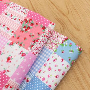 Può includere: Due pezzi di tessuto piegati con un design patchwork. I tessuti presentano un mix di quadrati rosa, bianchi e blu con motivi floreali e a pois. I tessuti sono probabilmente per il quilting o l'artigianato.