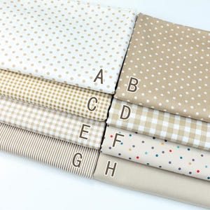 Puede incluir: Una selección de ocho muestras de tela en varios patrones y colores. Los patrones incluyen lunares, cuadros, rayas y colores lisos. La paleta de colores es principalmente neutra, con tonos beige, marrón claro y blanco. Cada muestra está etiquetada con una letra.