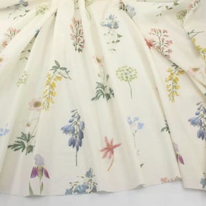 Può includere: Tessuto bianco sporco con un motivo floreale. Il design presenta vari fiori nei toni pastello di rosa, viola, giallo e blu, sparsi sul tessuto. Il tessuto è drappeggiato, mostrando pieghe e consistenza.
