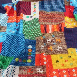 Op de afbeelding: Een patchwork textiel met een levendige mix van kleuren en patronen. Het ontwerp bevat vierkanten van blauwe, rode, oranje, groene en gele stof. Elk vierkant heeft een uniek patroon, waaronder zigzags, ruiten en bloemmotieven. Het algehele effect is een kleurrijk en gestructureerd ontwerp.