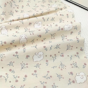 May include: Cream-coloured fabric featuring a repeating pattern of white bunnies, pink roses, and grey sprigs. The fabric is folded, displaying the pattern's continuity. Ideal for sewing, quilting, or crafting projects.