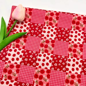 May include: A patchwork fabric featuring a red and white apple design, with gingham and floral squares. A pink tulip and green leaves are in the upper left. The fabric is suitable for quilting or crafting projects.