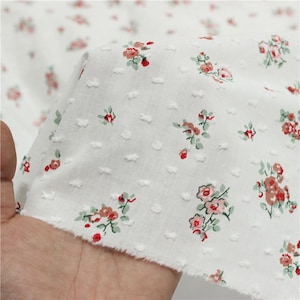 May include: White fabric with a delicate floral pattern featuring small pink and red flowers with green leaves. The fabric has a textured, dotted surface. The fabric is being held up, showing the raw edge.