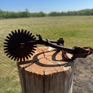 May include: A rusty metal spur with a star-shaped rowel rests on a weathered wooden post. The spur features a curved shank and leather strap, set against a backdrop of a green field and blue sky.