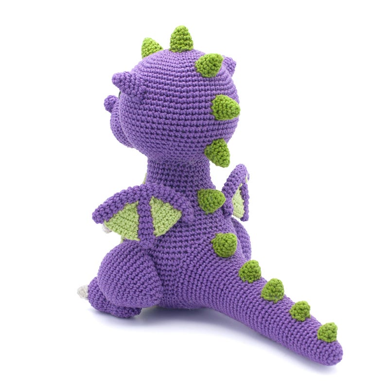 May include: Purple crocheted dragon toy with green accents. The dragon has a long tail, wings, and a spiky head.