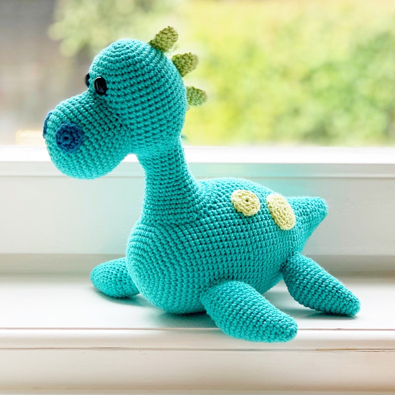 May include: A turquoise crocheted dinosaur toy with yellow spots. The dinosaur is lying on a white surface.
