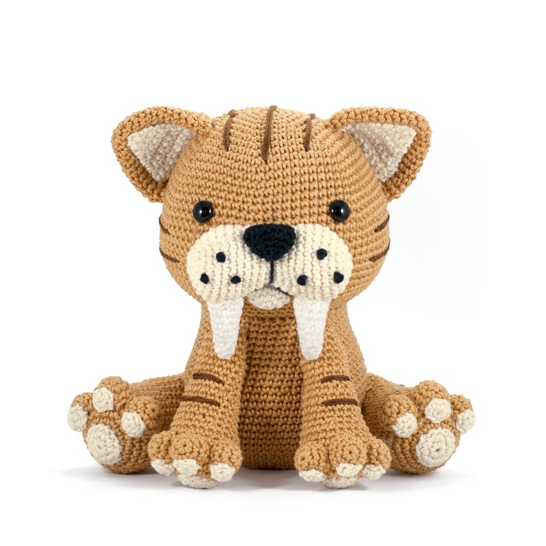 May include: A crocheted stuffed toy saber-toothed cat with brown fur, white fangs, and black eyes. The cat is sitting with its paws outstretched.