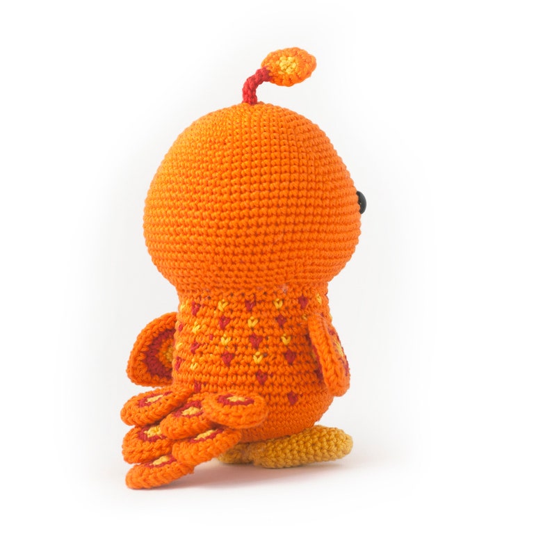 May include: Orange crocheted bird toy with a yellow beak and red and yellow tail feathers. The bird is standing on a white surface.