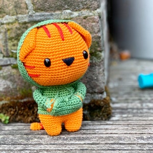 Pode incluir: Um brinquedo de tigre laranja feito em croch&ecirc; com olhos de pl&aacute;stico preto e nariz bordado em preto. O tigre apresenta listras vermelhas na cabe&ccedil;a e veste um moletom verde com capuz, bolso frontal e capuz combinando sobre as orelhas.