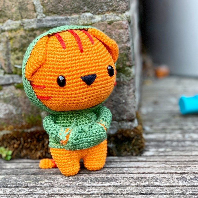 Pode incluir: Um brinquedo de tigre laranja feito em croch&ecirc; com olhos de pl&aacute;stico preto e nariz bordado em preto. O tigre apresenta listras vermelhas na cabe&ccedil;a e veste um moletom verde com capuz, bolso frontal e capuz combinando sobre as orelhas.