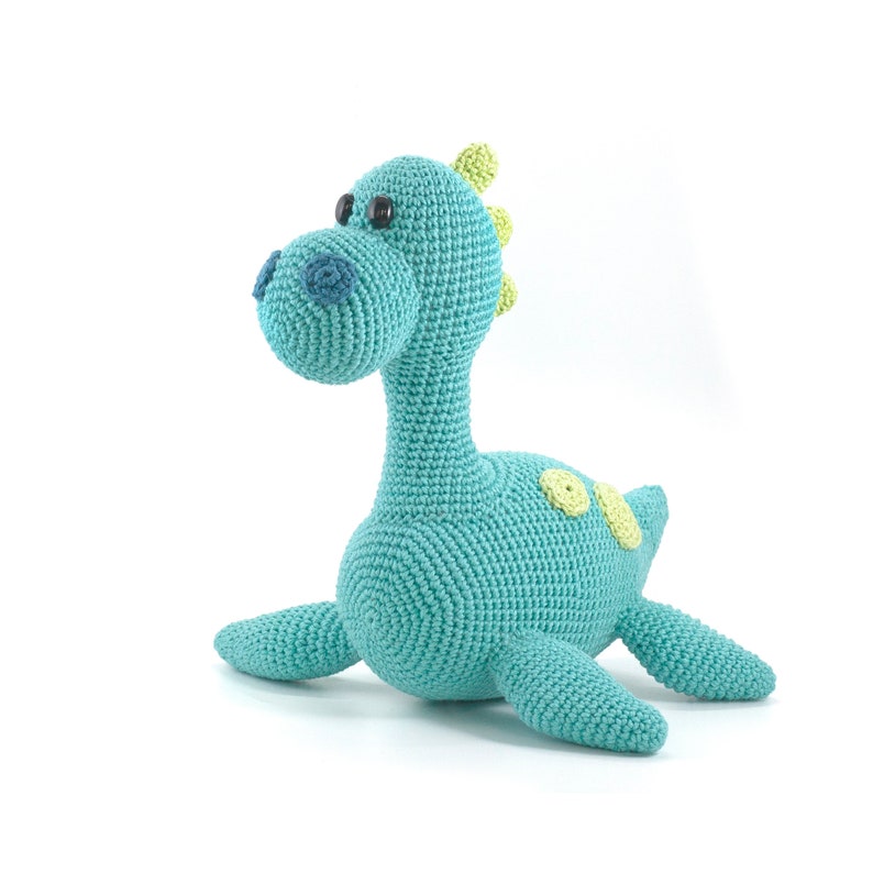 May include: A crocheted turquoise dinosaur toy with green accents. The dinosaur has large black eyes and a small blue nose.