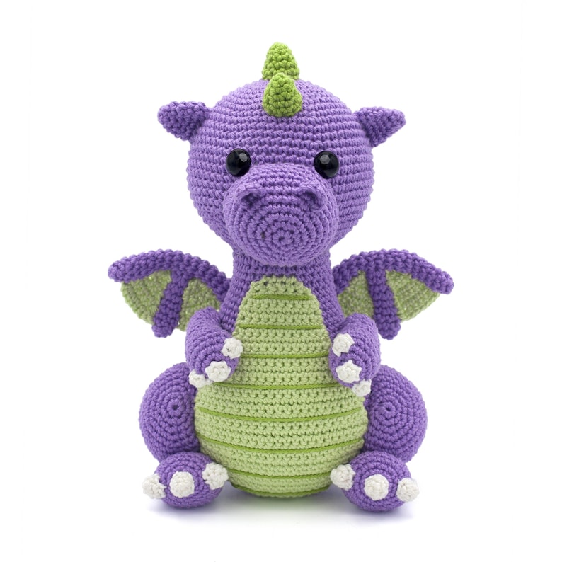 May include: A purple and green crocheted dragon toy with white claws. The dragon has a green horn and black eyes.