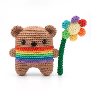 May include: A crocheted brown bear with a rainbow stripe across its chest, holding a colorful crocheted flower. The bear has black button eyes and a black nose. The flower has a green stem and a multi-colored flower head.
