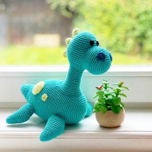 May include: A turquoise crocheted dinosaur toy with yellow spots. The dinosaur is sitting on a white surface next to a small potted plant.