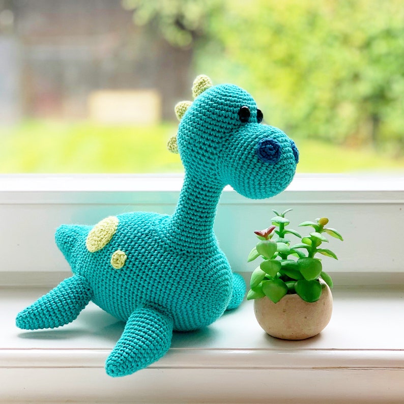 May include: A turquoise crocheted dinosaur toy with yellow spots. The dinosaur is sitting on a white surface next to a small potted plant.