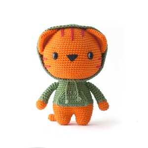 Pode incluir: Um peluche de tigre laranja feito em croch&eacute;, vestindo uma camisola verde com capuz. O tigre tem olhos de pl&aacute;stico preto, um nariz triangular preto e riscas vermelhas bordadas na testa e nas bochechas. A camisola verde tem um bolso frontal.