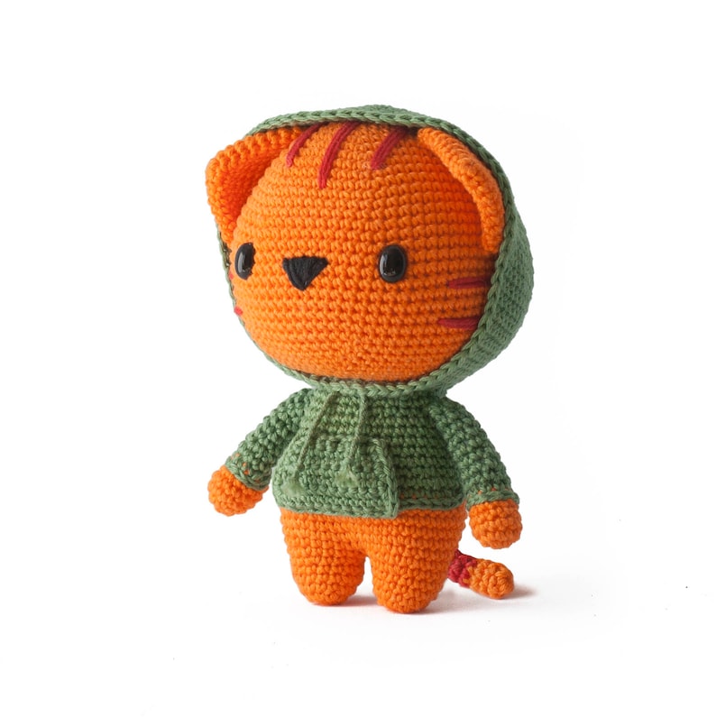 Pode incluir: Um bicho de pel&uacute;cia feito em croch&ecirc; com formato de tigre laranja com listras vermelho-escuras, olhos de pl&aacute;stico preto e nariz preto. O tigre veste um moletom com capuz verde-s&aacute;lvia com bolso frontal e cord&otilde;es combinando.
