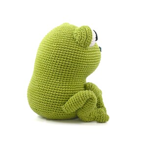 May include: A handmade, green crocheted frog doll. The frog is sitting with its legs bent, and its eyes are black and white. The frog is made of a soft, textured yarn and is a cute, whimsical toy.