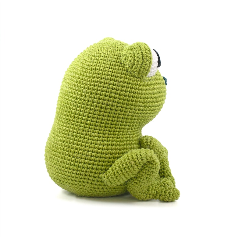 May include: A handmade, green crocheted frog doll. The frog is sitting with its legs bent, and its eyes are black and white. The frog is made of a soft, textured yarn and is a cute, whimsical toy.