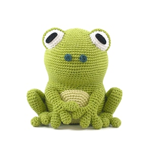 May include: A handmade crochet frog in a sitting position. The frog is primarily light green with a beige belly and feet. It has large white-rimmed eyes with black centers and small blue eyes. The frog's arms are crossed, and it has a textured, stitched appearance.