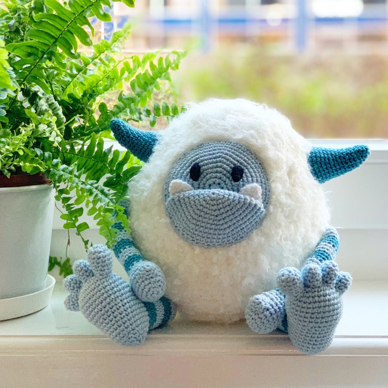 May include: A crocheted plush toy resembling a yeti or abominable snowman. The toy is white with blue and turquoise accents. It has a fluffy white body, blue crocheted arms and legs, and a blue crocheted face with black eyes.