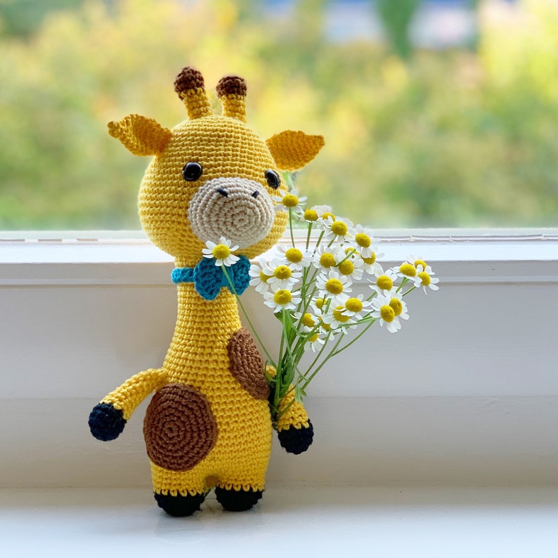 May include: A crocheted giraffe toy holding a bouquet of white daisies. The giraffe is yellow with brown spots and a blue bow tie.