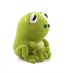 May include: A handmade, green crochet frog plush toy. The frog has large white eyes with black pupils, a light tan belly, and small blue-green nostrils. The frog is sitting with its arms crossed and legs tucked, showcasing the detailed crochet work.