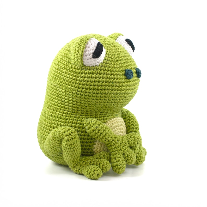 May include: A handmade, green crochet frog plush toy. The frog has large white eyes with black pupils, a light tan belly, and small blue-green nostrils. The frog is sitting with its arms crossed and legs tucked, showcasing the detailed crochet work.