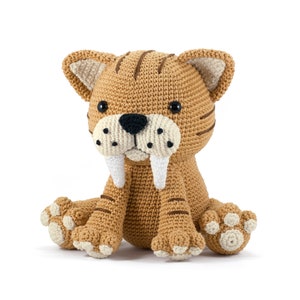 May include: A crocheted stuffed toy saber-toothed cat with brown fur, white fangs, and black eyes. The cat is sitting with its paws outstretched.