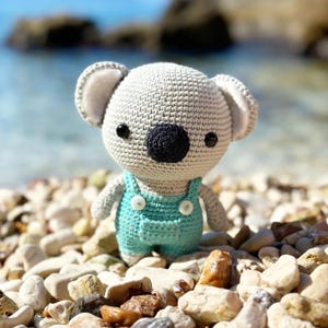 May include: A crocheted koala bear doll with a gray body, black eyes, and a black nose. The koala wears light blue overalls with white buttons. The doll is standing on a bed of pebbles near the ocean.