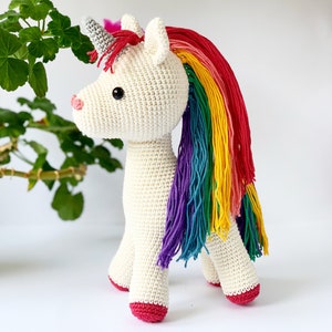 May include: A crocheted unicorn toy with a cream-colored body, a gray horn, and a rainbow mane. The unicorn has a pink nose and red hooves. The toy is standing on a white surface.