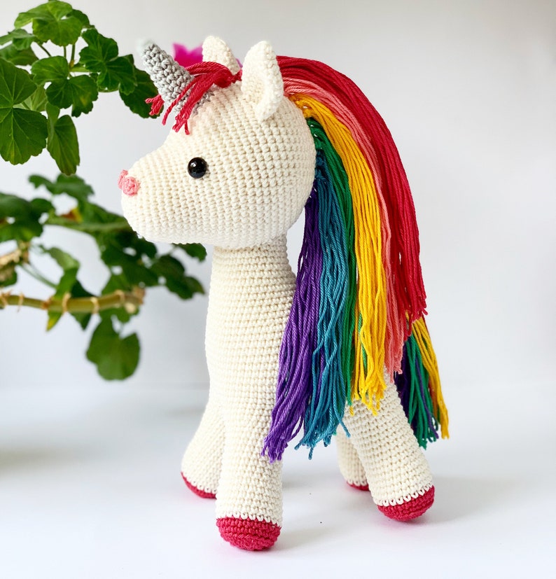 May include: A crocheted unicorn toy with a cream-colored body, a gray horn, and a rainbow mane. The unicorn has a pink nose and red hooves. The toy is standing on a white surface.
