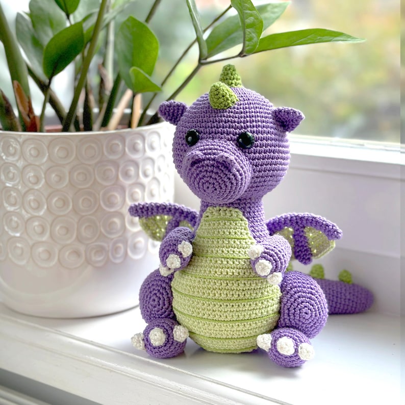 May include: A crocheted purple and green dragon toy with white accents. The dragon is sitting on a windowsill.