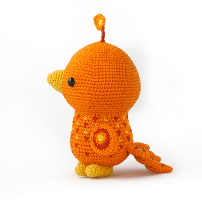 May include: A crocheted orange bird with a yellow beak and feet. The bird has a small orange crest on its head and a patterned orange, yellow, and red tail.