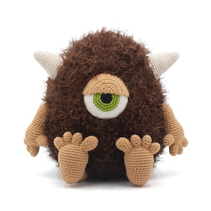 May include: A brown crocheted monster with a single green eye, white horns, and brown feet.