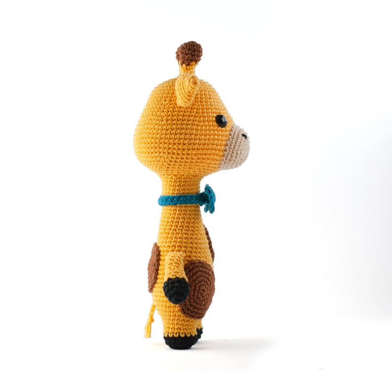 May include: A crocheted giraffe toy with brown spots and a blue bow tie. The giraffe is standing on a white surface.